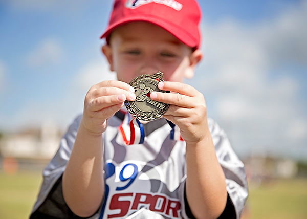 a pee wee age group baseball player in red hat and grey jersey shows off his sportsmanship medal holding it in two hands.
