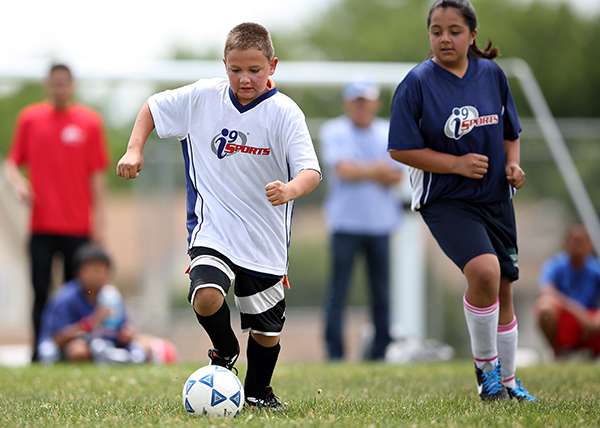 Youth Soccer player in white i9 Sports jersey dribbles a soccer ball during a kids soccer game as a defender in a blue jersey approaches.