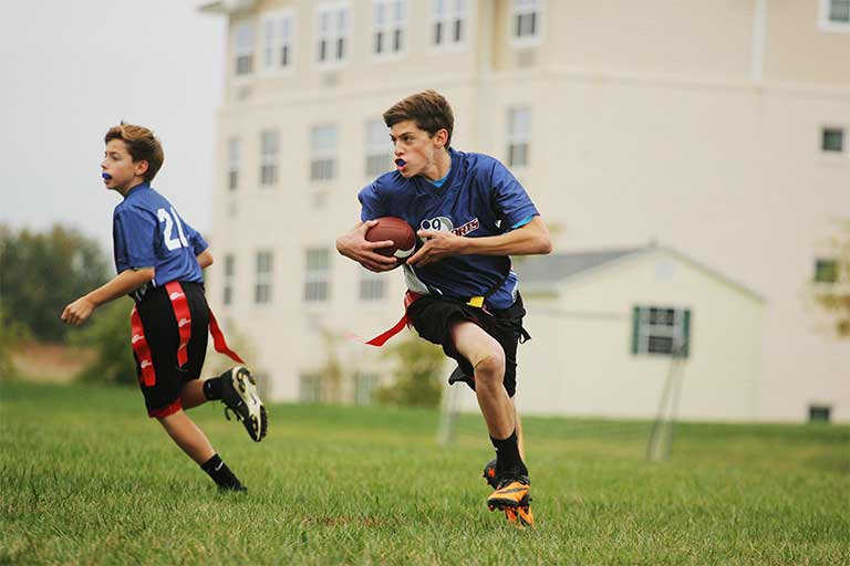 Male flag football player in blue i9 Sports jersey runs with the football tucked under his right arm.