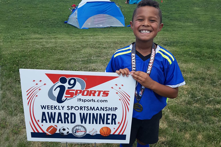 Youth soccer player smiles and poses with i9 Sports weekly sportsmanship award winner sign while wearing his sportsmanship medalship medal.