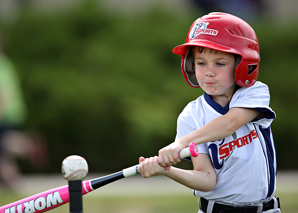 i9 Sports youth t-ball player hits a baseball off a tee wearing a white i9 Sports jersey and red batting helmet