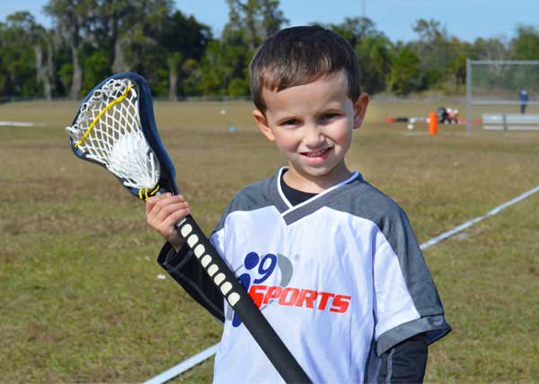Smiling boy in white i9 Sports jersey holds a lacrosse stick