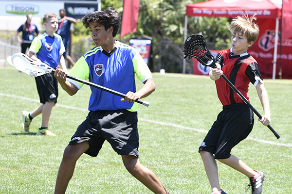 A lacrosse player in an i9 Sports jersey catches the lacrosse ball as a defender in a red jersey attempts to intercept the ball