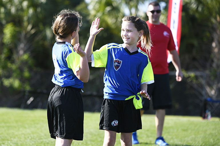 i9 Sports soccer player gives a high five to a team mate while coach looks on from sideline.