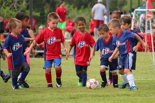 A group of youth soccer players wearing red and blue i9 Sports soccer jerseys crowd around the soccer ball trying to kick it.