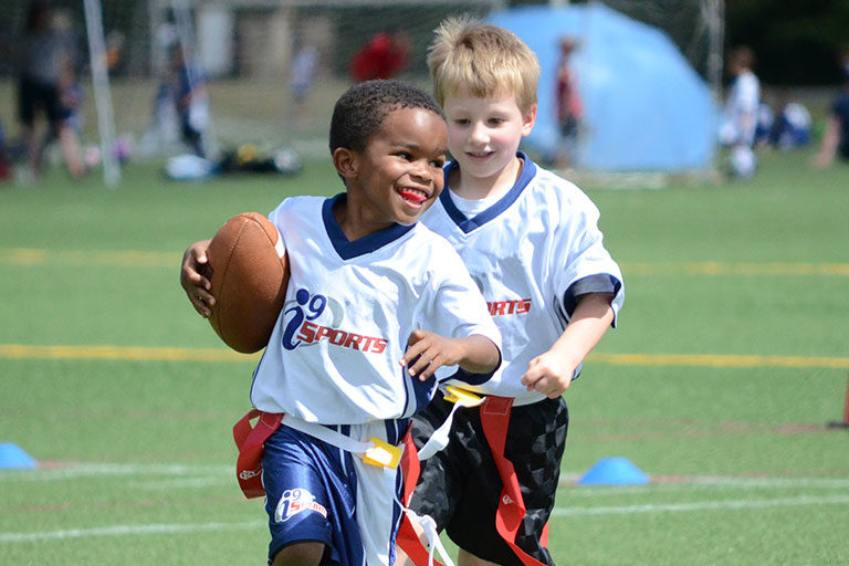 Young flag football player carries the football while running toward the end zone as a team mate looks on.