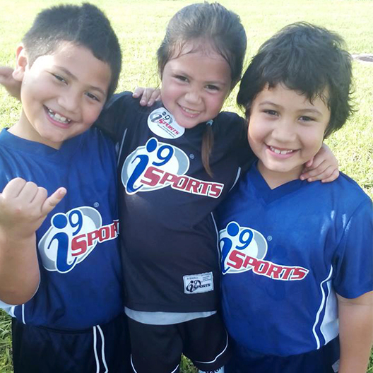 Three kids wearing i9 Sports soccer jerseys huddle together smiling after their soccer game in Honolulu