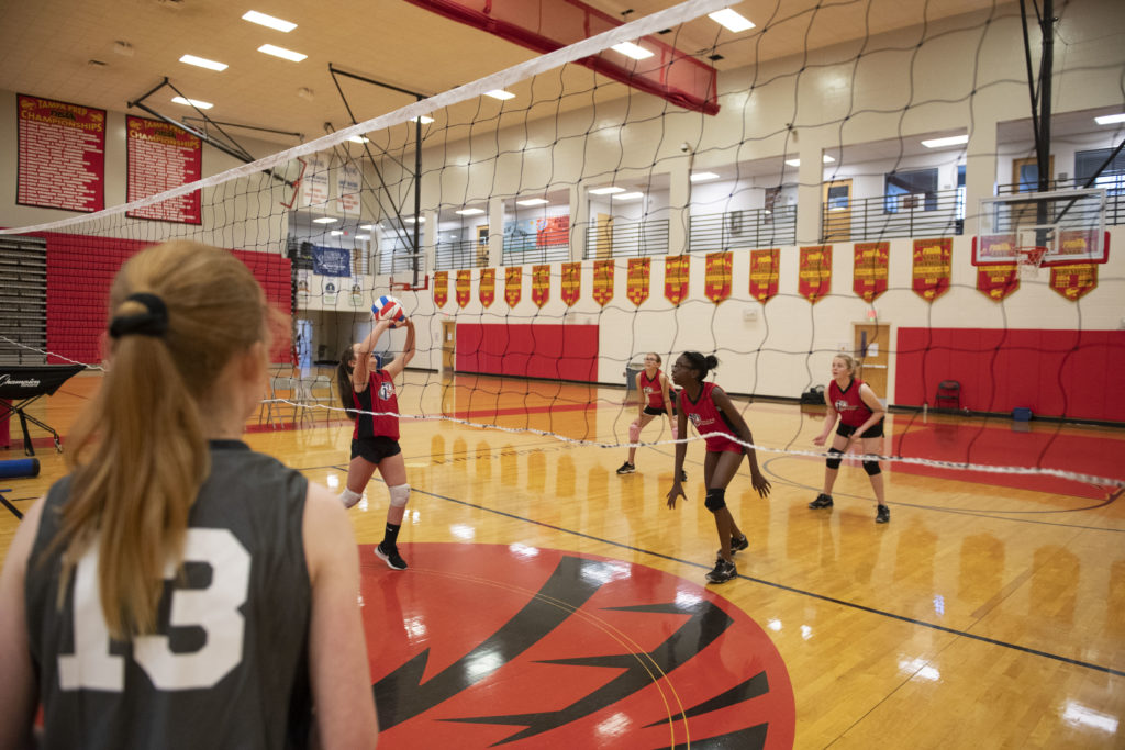 Teenage volleyball player in a gray jersey with the number 13 looking through the net at the other team in red jerseys passing the volleyball.