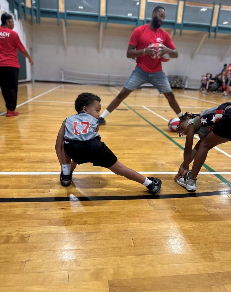 Little boy in a gray jersey with the number 17 at basketball practice lunging to his left side to stretch his right leg.
