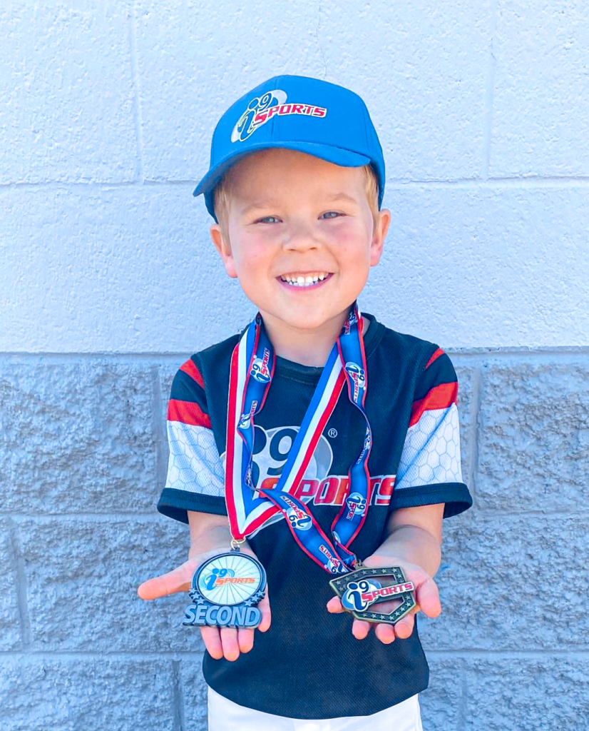 Young boy in a blue i9 Sports baseball hat and black i9 Sports baseball jersey holding his sportsmanship medal on the right and his second place medal on the left.