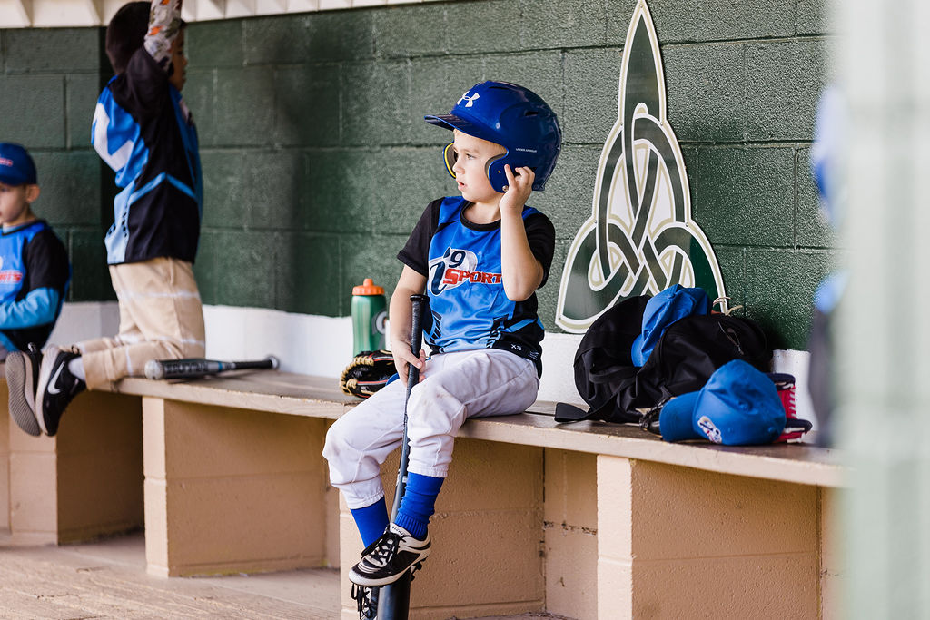 A young boy in a blue baseball helmet and blue i9 Sports jersey is sitting in the dugout holding his bat.