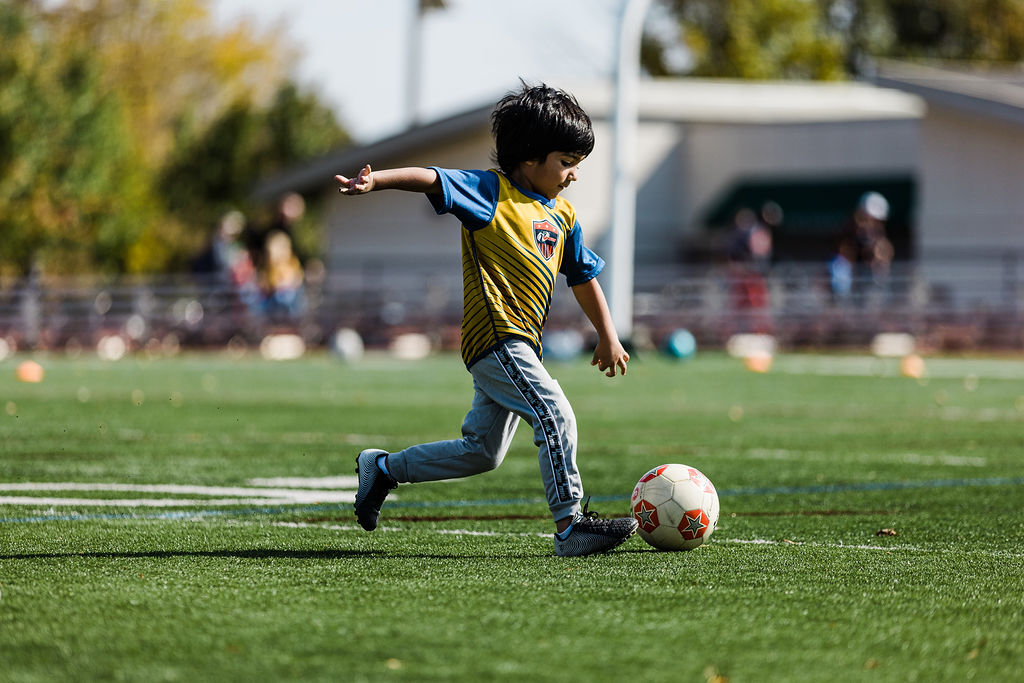 Young boy in an i9 Sports soccer jersey at soccer practice kicking a soccer ball.