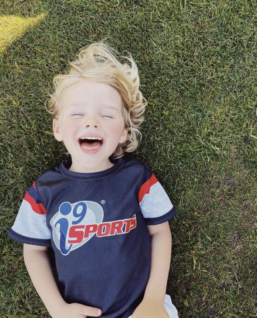 Young blond haired child laying in the grass laughing with eyes closed.
