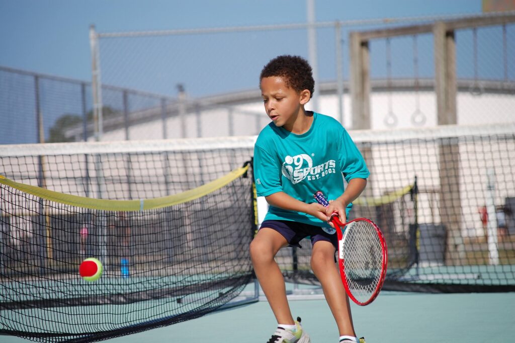 Young boy in an i9 Sports tennis shirt playing tennis on a green tennis court.