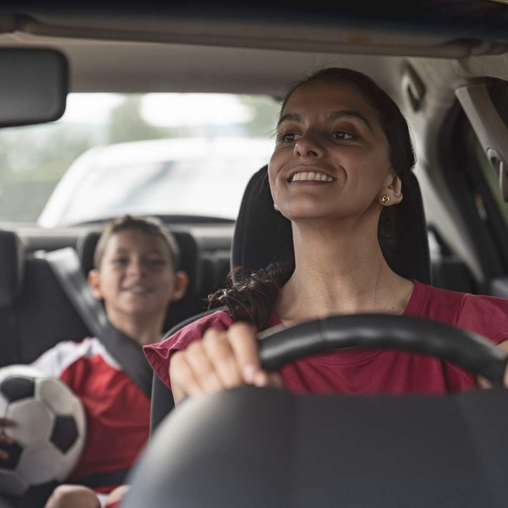 A mom in the drivers seat on a car, driving her son to soccer as he holds a soccer ball in the back seat.