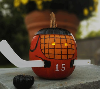 Pumpkin painted like a hockey player with a small hockey stick poking through it. Next to the pumpkin, is a baby pumpkin painted like a hockey puck.