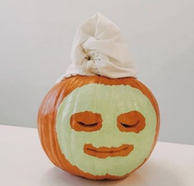 pumpkin painted like it's enjoying a spa day. It has a green face mask and a towel wrapped around the top.