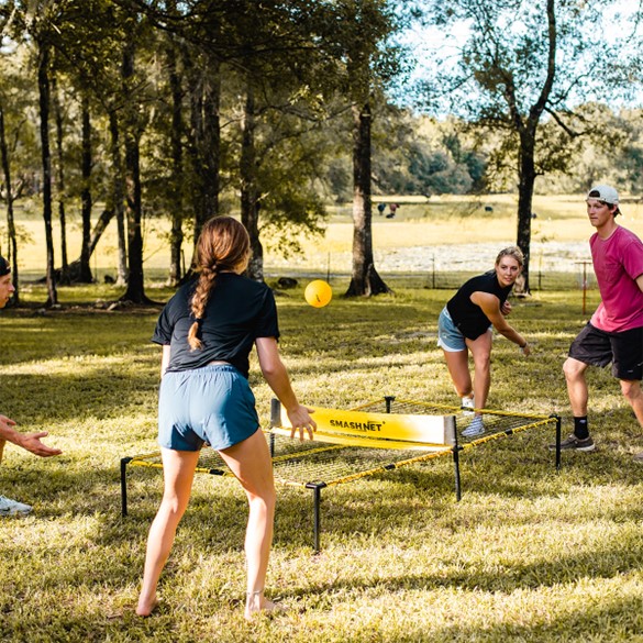 Young adults playing a game called SmashNet. Two players are on each side of a net that is sitting out the ground. They bounce a bright yellow ball onto the bouncing part of the play area and over the net to the other side. It's like a mini version of pickleball without the paddles.