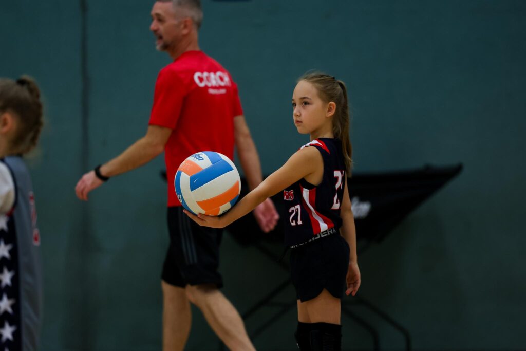 Young female athlete in a navy blue i9 Sports jersey holding a volleyball she is about to serve during a volleyball game.