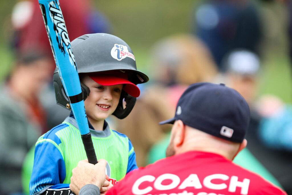 Young boy in an i9 Sports baseball helmet holding a bright blue baseball bat while a coach in a red shirt that says COACH helps put the boy's hands in the correct position.