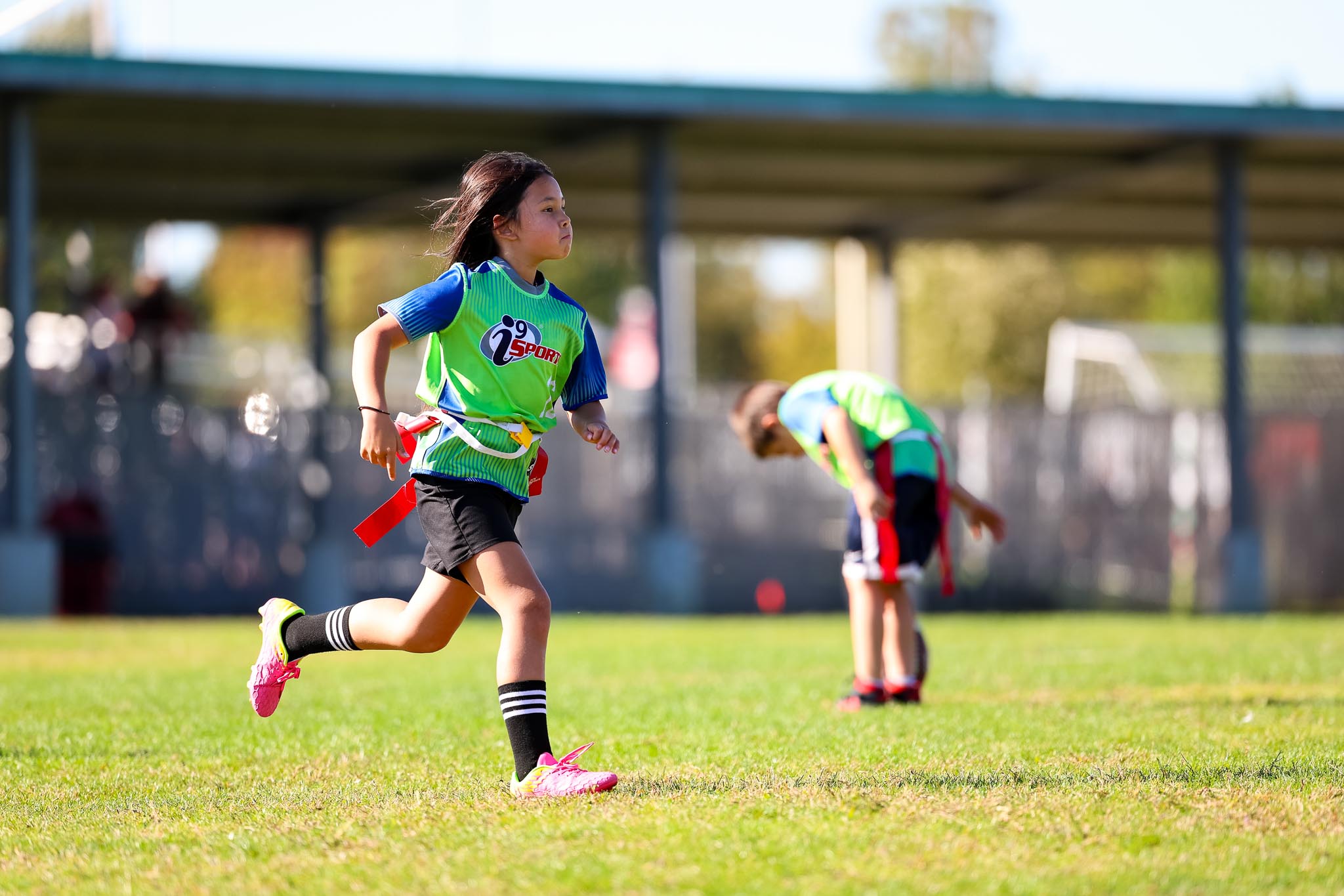 Girl in a bright green and blue i9 Sports flag football jersey running down the football field with her red flags on.