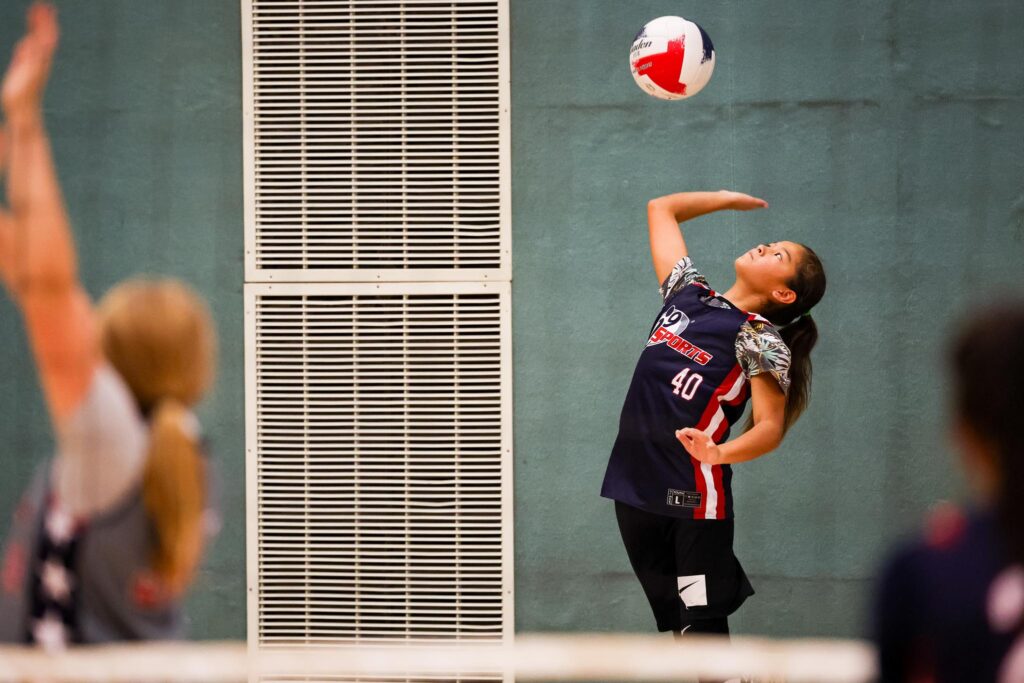 Girl in a navy blue i9 Sports volleyball jersey jumping up and hitting a red, white, and blue volleyball to serve the ball over the net to her opponents.
