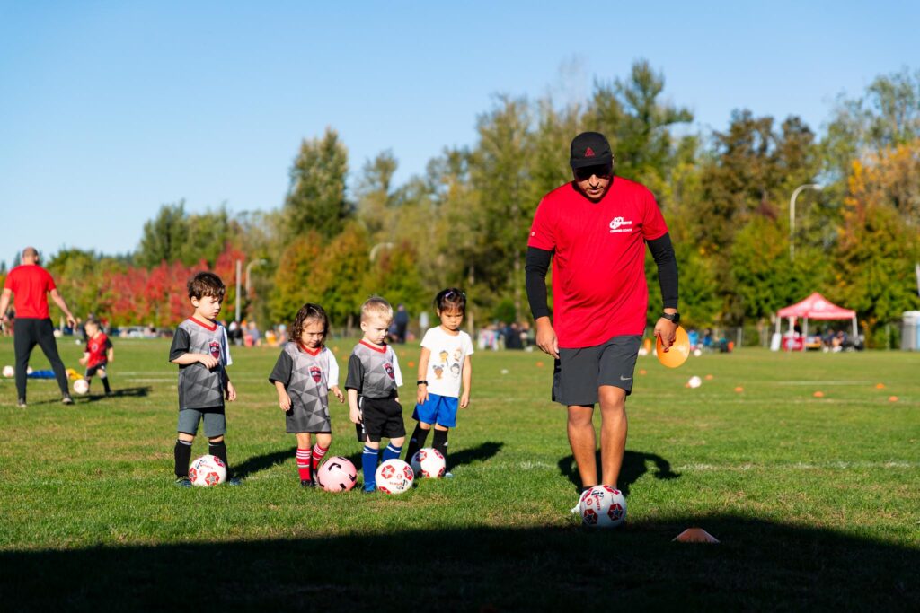 Four toddler aged kids in gray and red i9 Sports soccer jerseys following their coach while each of them kick their soccer ball.