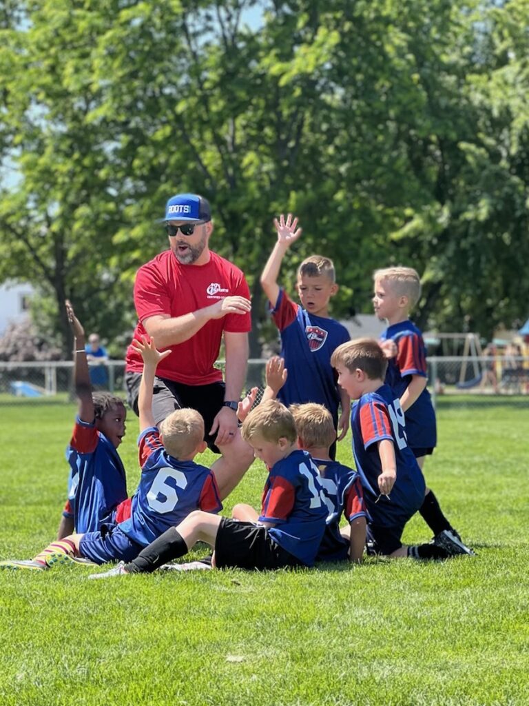 Coach in a red shirt kneeling on the ground while surrounded by his young soccer players in bright blue i9 Sports soccer jerseys all raising their hands in the air.