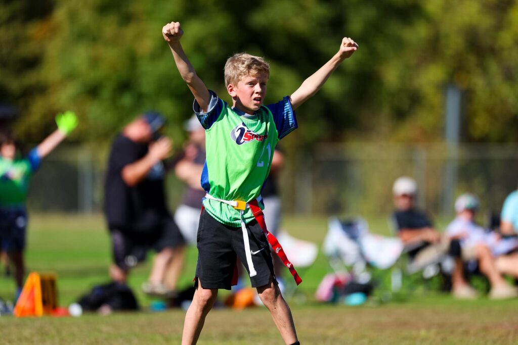 Pre-teen boy with blond hair in a bright green and blue i9 Sports jersey and flag football flags with both arms in the air celebrating the last play during an i9 Sports flag football game.