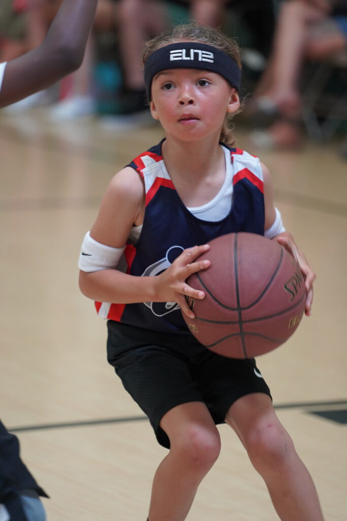 Young basketball player in a navy blue headband and navy blue i9 Sports jersey holding a basketball and about to pass it during a game.