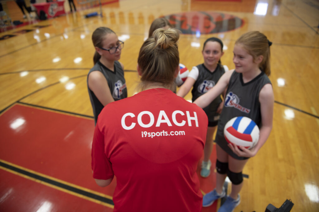 Girls volleyball team huddled up with their hands in. The coach has her back toward the camera so you can read COACH on the back of her bright red shirt.