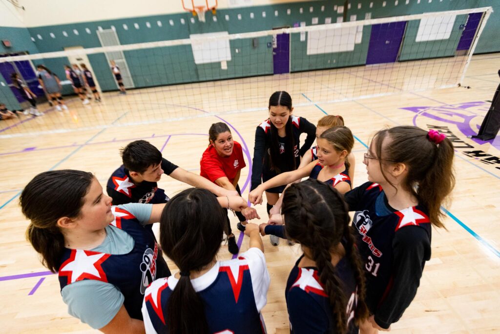 Volleyball players with the fists in the middle of a huddle with their female coach in a red shirt.