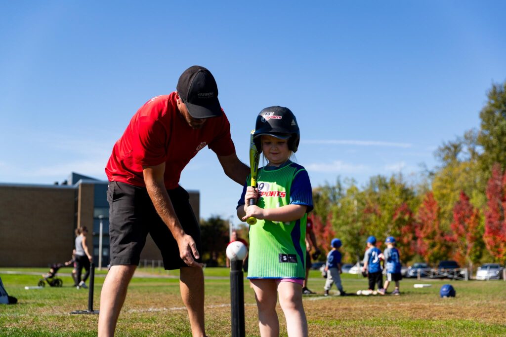 A coach on the left in a red shirt helping a young girl playing tball. She is in a bright green and blue i9 Sports jersey with a bright yellowish green bat on her left shoulder. She is standing over the tee and ball while the coach shows her where to stand.
