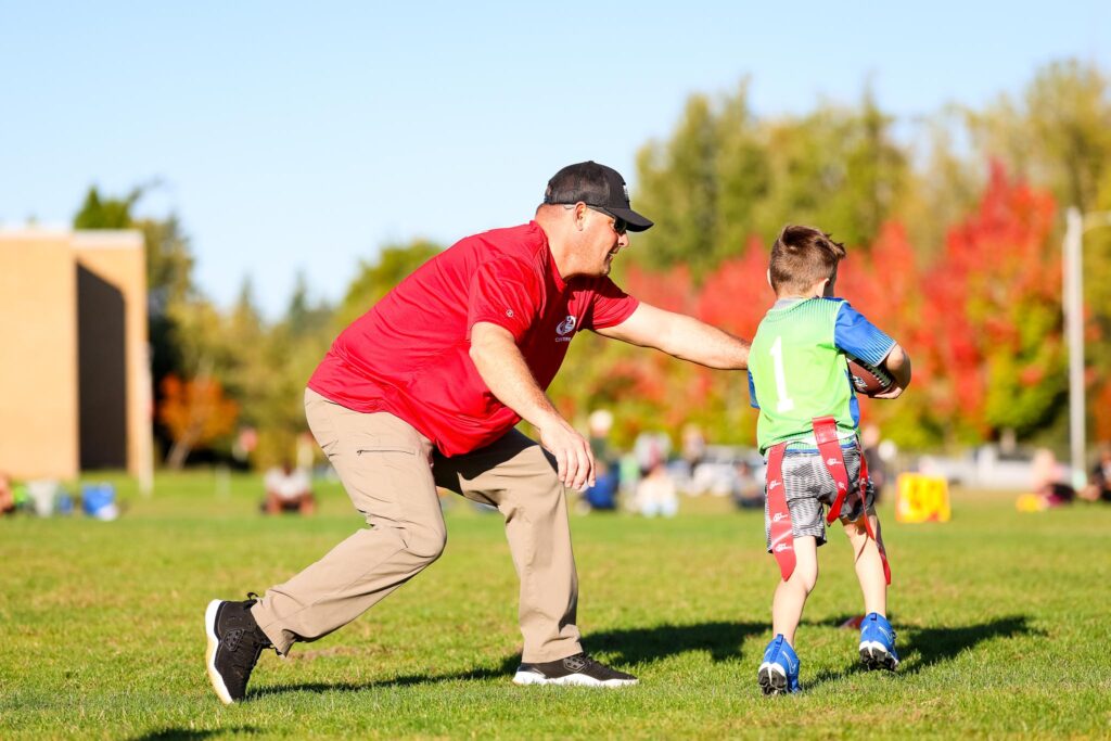 The coach in a red shirt on the left is handing the football off to his young flag football player to practice receiving hand offs.