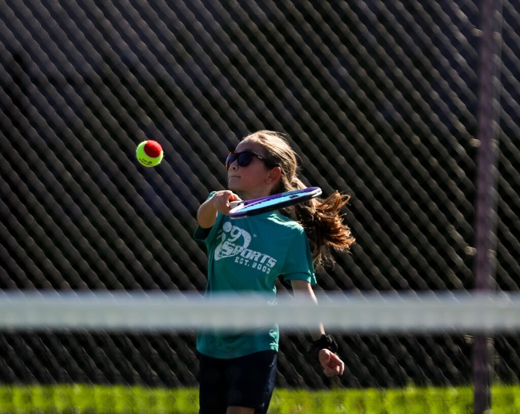 Young girl playing tennis in a teal i9 Sports t-shirt. She is wearing sunglasses and swinging a tennis racket with her right hand at a bright red and green tennis ball.