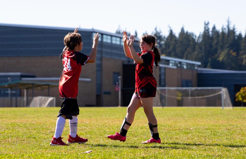 Boy soccer player on the left high fiving the girl soccer player on the right with both hands. Both are wearing red and black i9 Sports soccer jerseys.