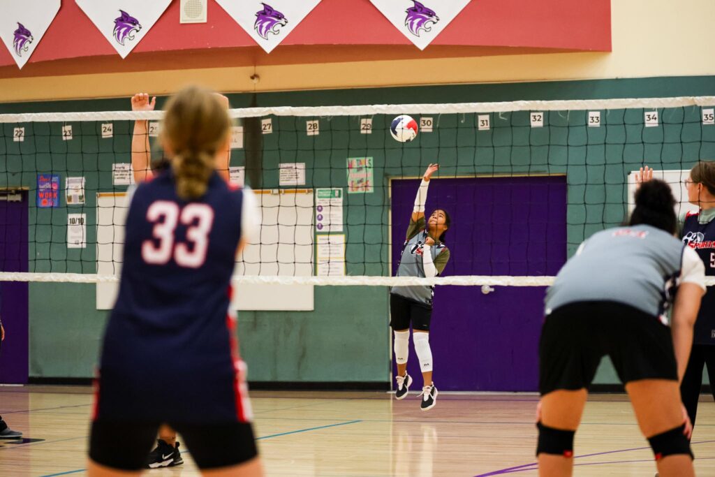 Teenage girls playing team volleyball indoors as one girl serves the ball overhand from the other side of the net.