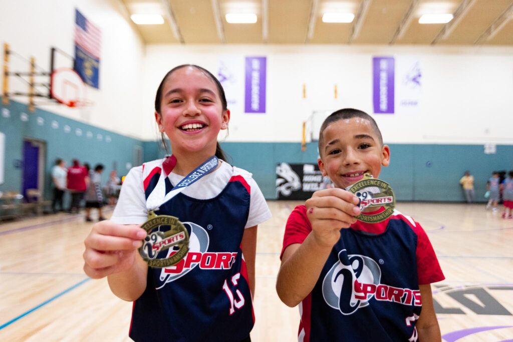 Two i9 Sports basketball players, girl on the left and boy on the right, holding up their sportsmanship medals that they won for showing an act of sportsmanship during their game.