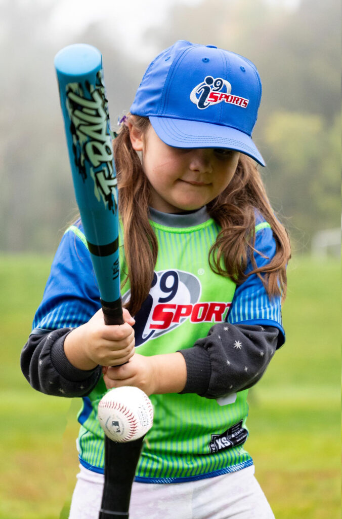 Young girl in a royal blue i9 Sports hat and bright green and blue i9 Sports jersey playing t-ball. She is lining up her bat with the ball on a tee.