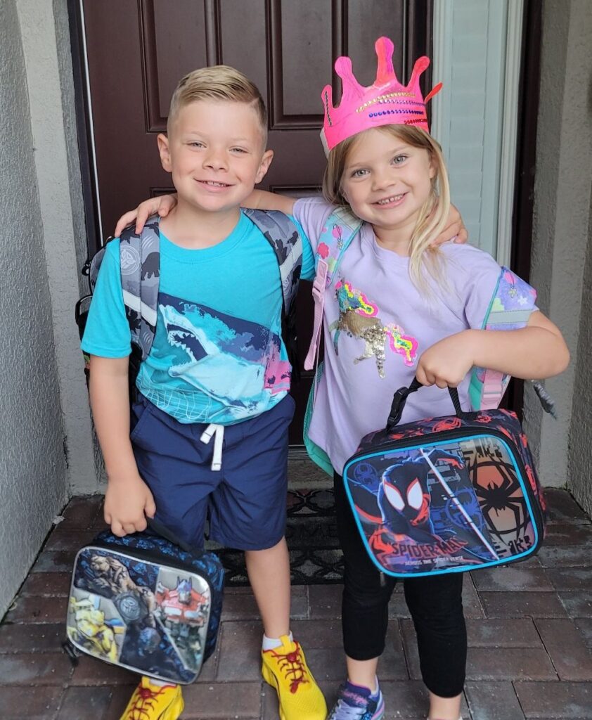 A brother and sister posing for a back to school photo. The boy on the left has blond hair and is wearing a bright blue shark t-shirt, navy blue shorts, and yellow shoes with red shoelaces. The girl on the right has longer blond hair and is wearing a pink paper crown, purple t-shirt, black leggings, and purple shoes. They both have lunchboxes with popular cartoons on them.