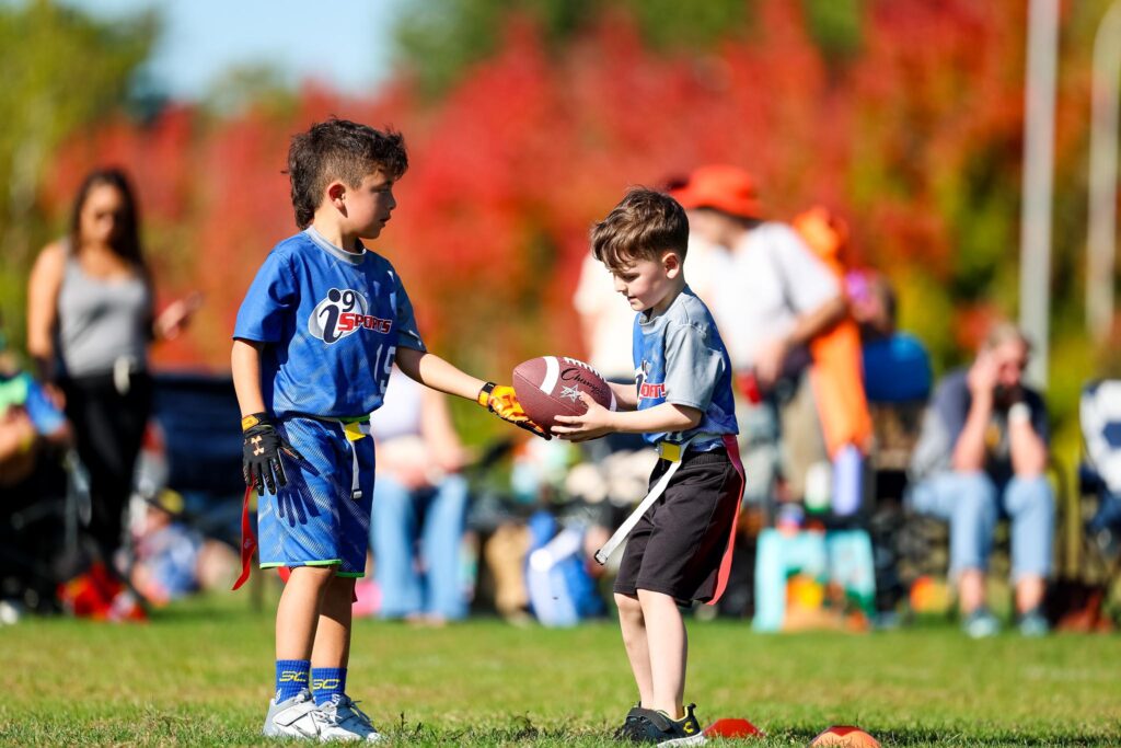 Two young flag football players in blue i9 Sports jerseys handing off a football during fall season with beautiful red trees in the background as the leaves change colors.