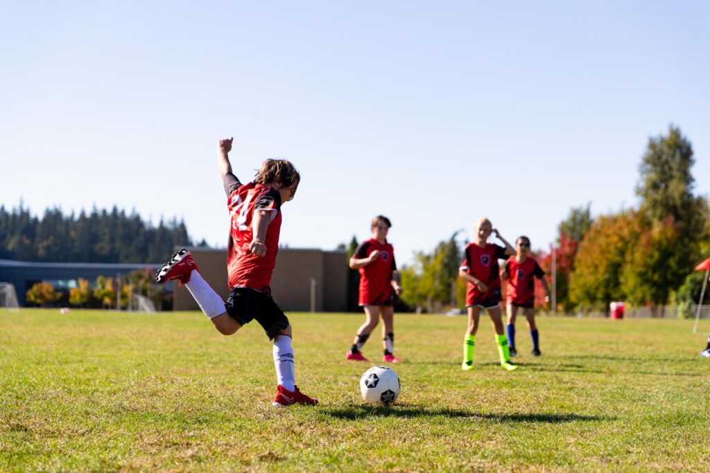Adolescent soccer player wearing a red and black jersey with the number 24 on the back. He has his left foot planted as he lifts his right leg backwards to kick a soccer ball with enough force to get it across the field.