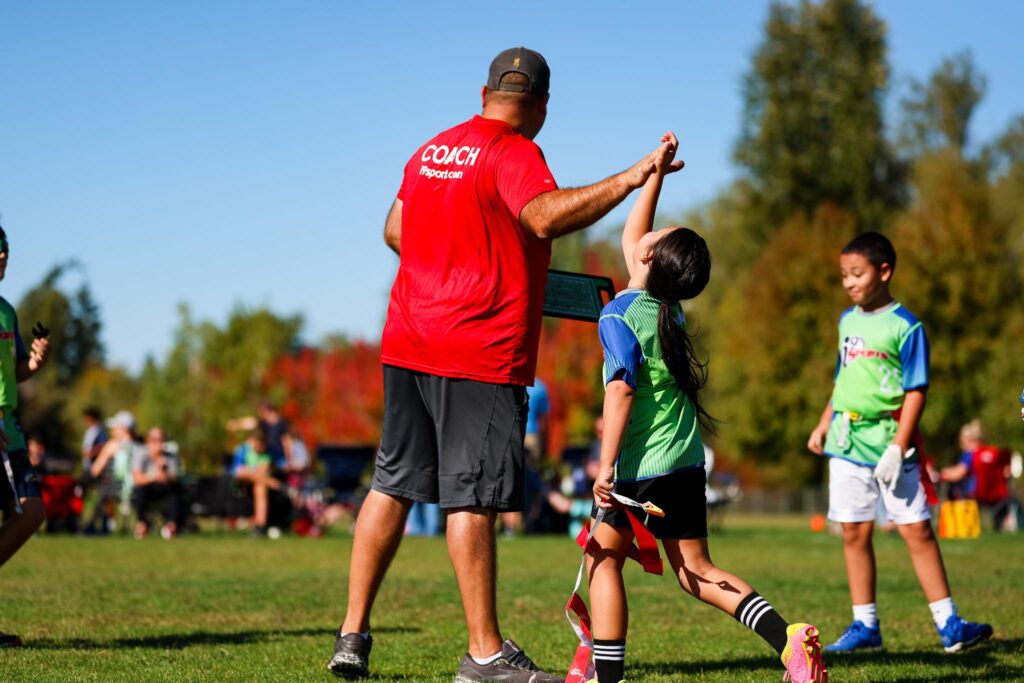 Flag football coach wearing a red shirt that says COACH on the back is high fiving the young female football player wearing a bright green and blue i9 Sports flag football jersey during their game.