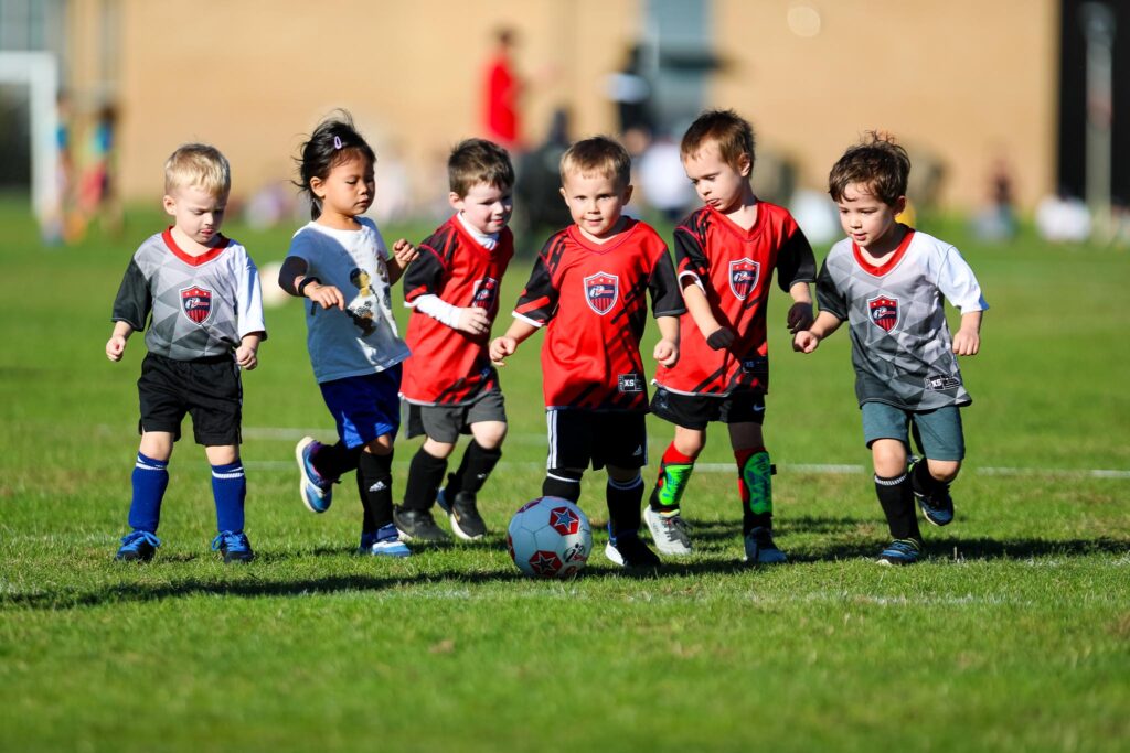 Six toddlers in a line running at the same soccer ball. All wearing red i9 Sports soccer jerseys or gray i9 Sports soccer jerseys.