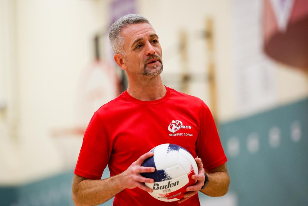 Volleyball coach in a red i9 Sports Coach shirts that reads i9 Sports Certified Coach on the right chest. He is holding a red white and blue volleyball.