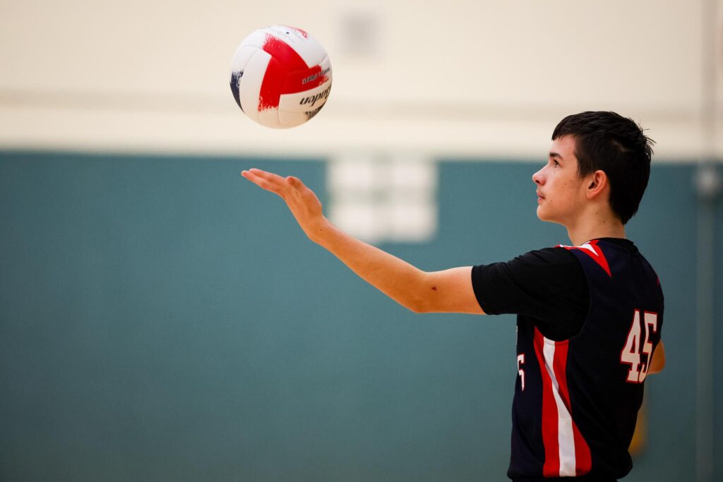 Adolescent boy with brown hair in a navy blue i9 Sports volleyball jersey standing with his side profile showing as he tosses a red white and blue volleyball in the air in front of him.