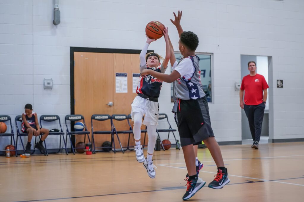 Smaller blond haired boy with a black sweat band on his head goes up for a jump shot against a larger adolescent player who is trying to block his shot.