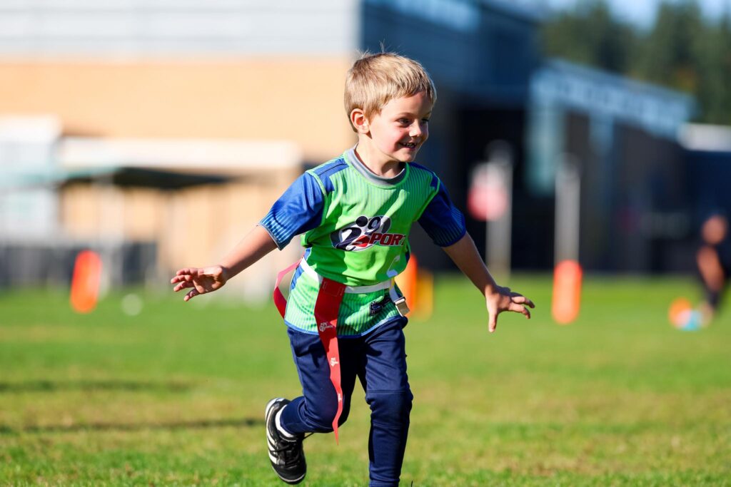 Young boy with blond hair wearing a bright green and blue i9 Sports jersey and flag football flags around his waist while he runs down the field with his arms spread open to each side like airplane wings.