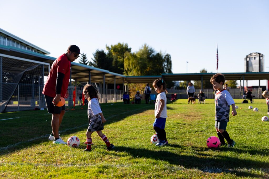 Young toddler-aged children lined up kicking soccer balls with their coach as they learn the basics of dribbling a soccer ball.