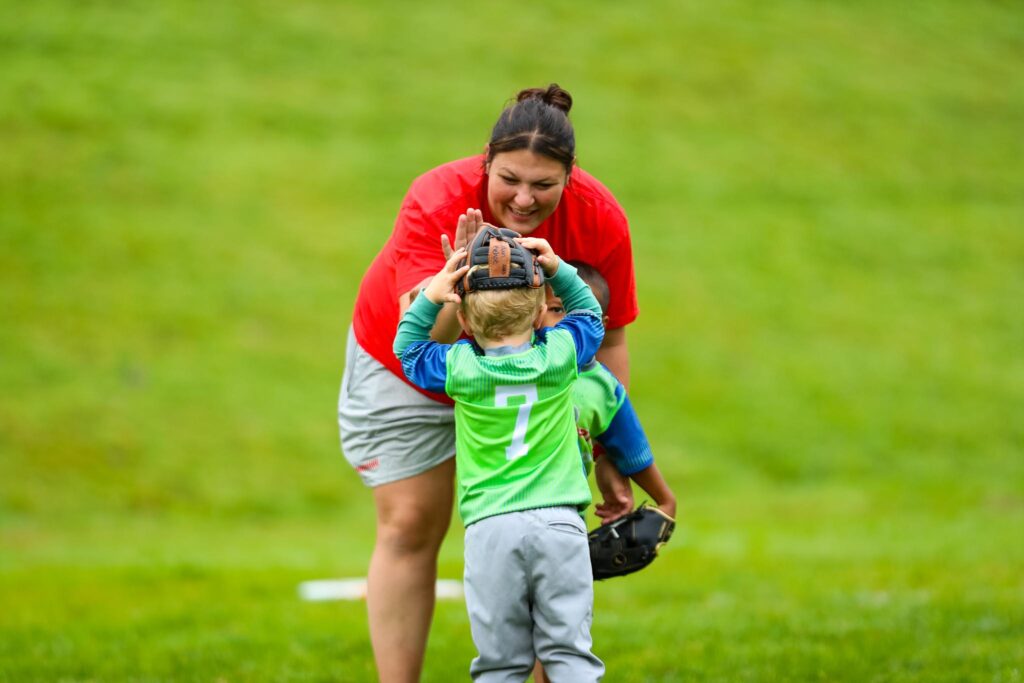 Woman coaching t-ball bends over to high five her t-ball player who is standing there holding a glove on his head. His back is facing the camera so you see his number 7 on the bright green jersey with royal blue short sleeves.